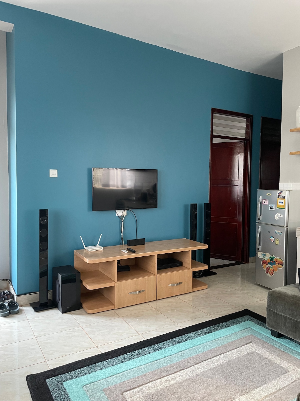 A living area is presented with a sleek, wooden entertainment unit and a flat-screen television mounted on a vibrant blue wall. Two tall speakers frame the unit, while a modern rug with subtle geometric patterns anchors the space. A refrigerator is positioned in the background.