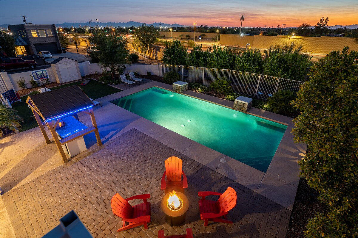 An aerial view of a heated pool area features a circular fire pit surrounded by bright red Adirondack chairs. The setting includes lounge chairs near the pool, with mountains visible in the distance under a twilight sky.