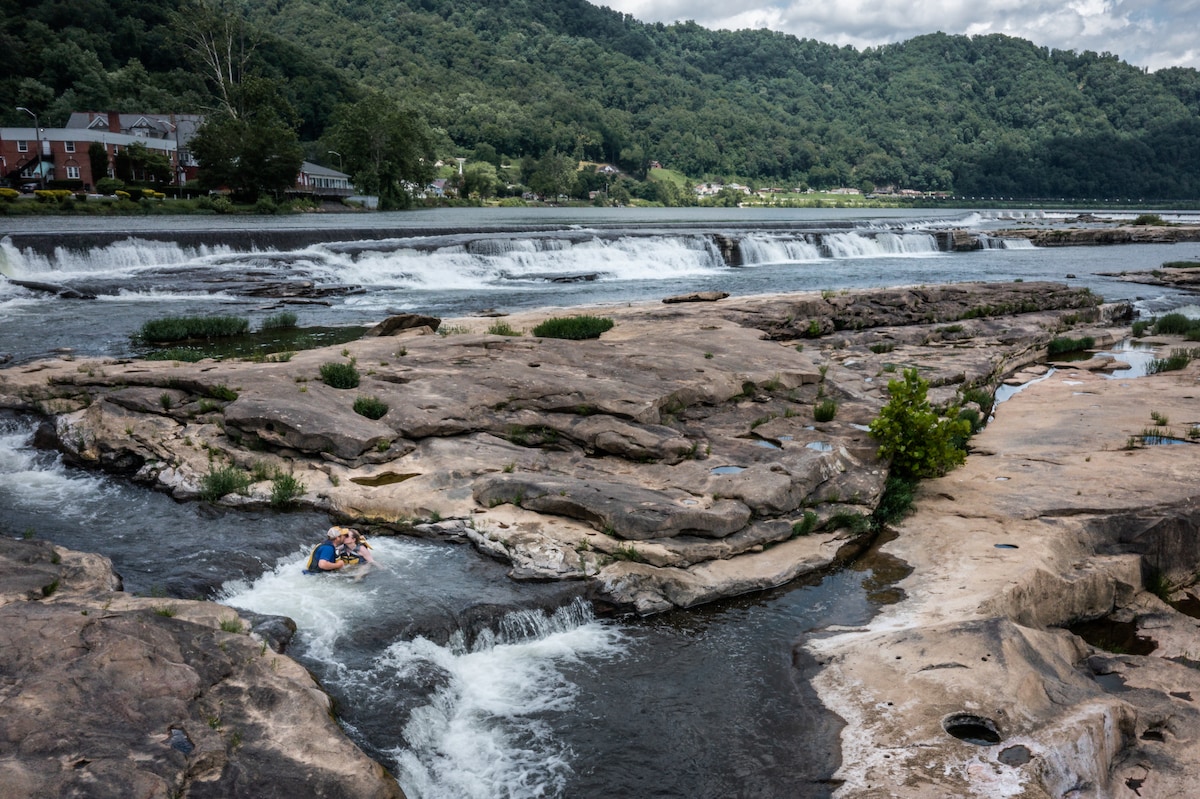 A wide view of a rocky river landscape is presented, with a gentle cascading waterfall in the background. Two individuals are seen floating on the water, surrounded by smooth rock formations and lush greenery along the riverbank. The mountains rise in the distance under a partly cloudy sky.