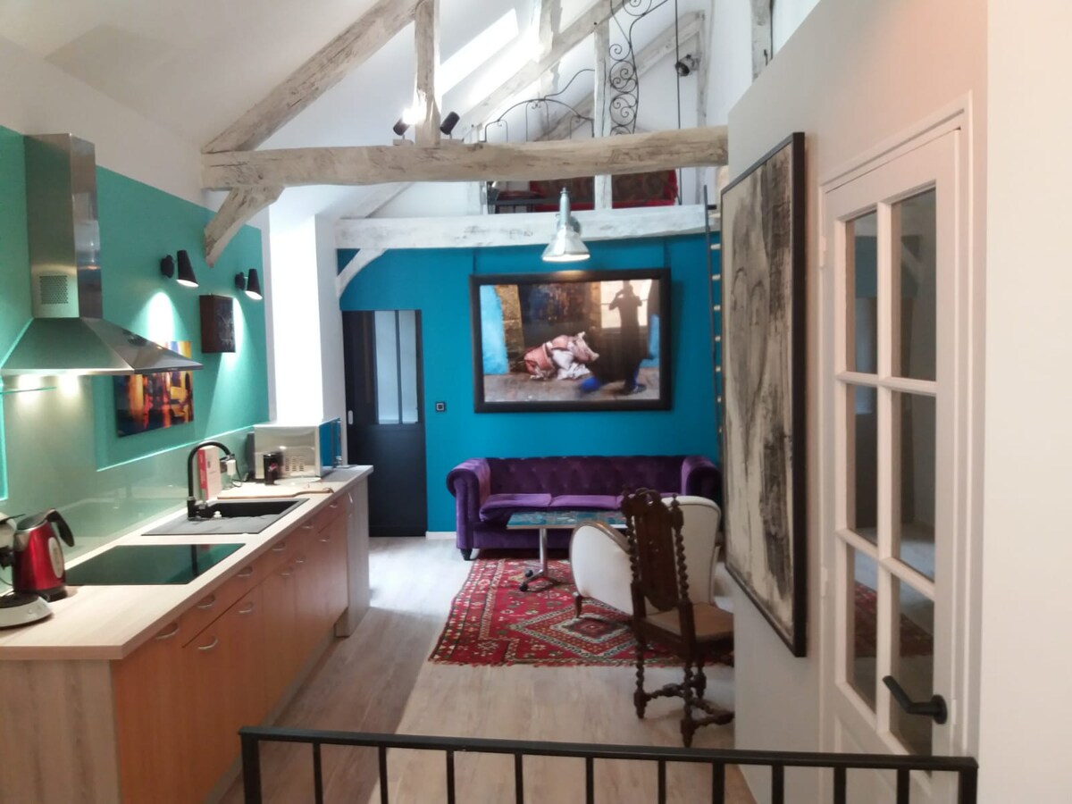 A bright, open space features a well-equipped kitchen with modern appliances and a stylish countertop. A purple sofa is positioned near a decorative chair, with an area rug providing warmth. Art pieces adorn the walls, and beams add character to the high ceilings.