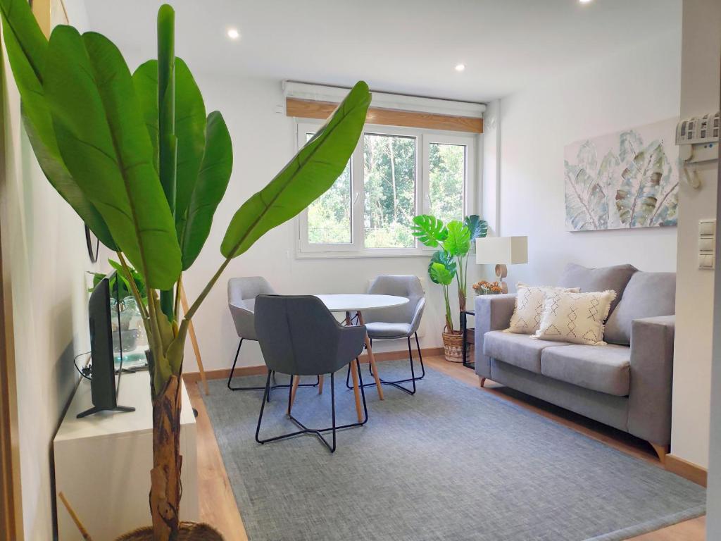 A light-filled living area features a comfortable gray sofa and a small dining table surrounded by four chairs. A large potted plant adds a touch of greenery, while a soft area rug complements the wooden flooring. Two windows allow natural light to stream in.