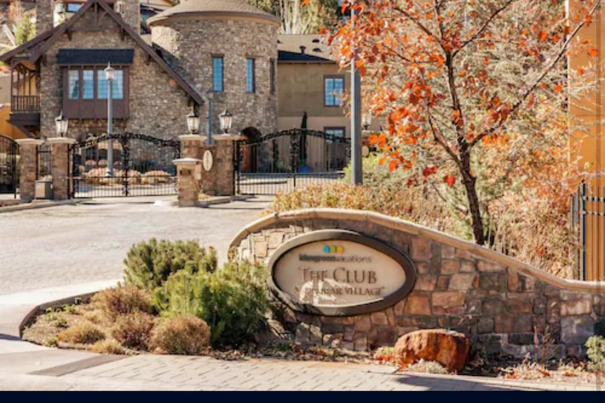 The entrance to the resort is marked by a stone sign displaying 'The Club at Big Bear Village.' Surrounding shrubs and seasonal foliage complement the welcoming gated access, while a distinctive stone structure can be seen in the background, adding character to the setting.