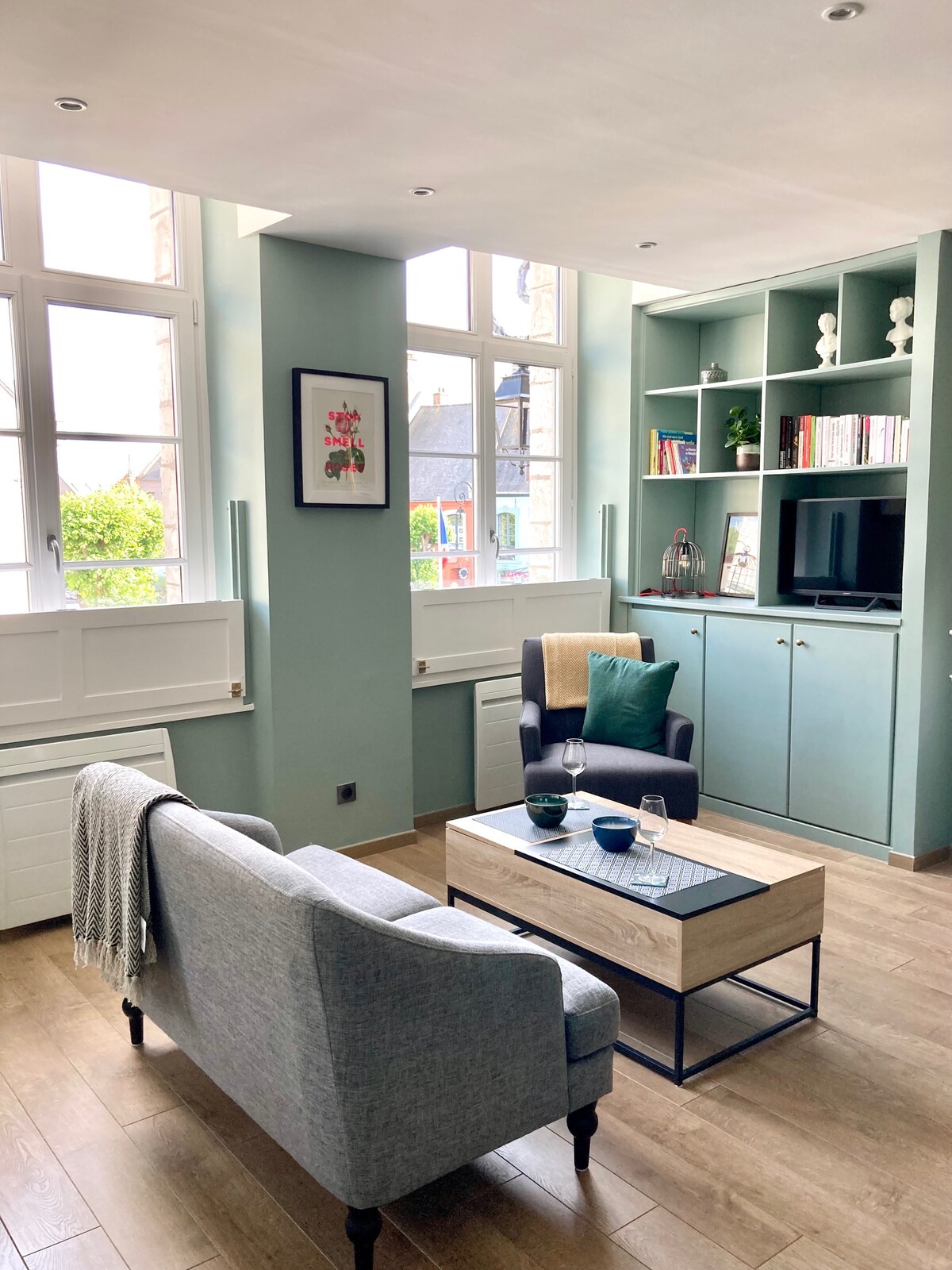 A bright living space is depicted, featuring a comfortable seating arrangement with a light gray sofa and a stylish armchair. A wooden coffee table occupies the center, complemented by decorative items. Large windows allow natural light to illuminate the area, enhancing the cozy ambiance.