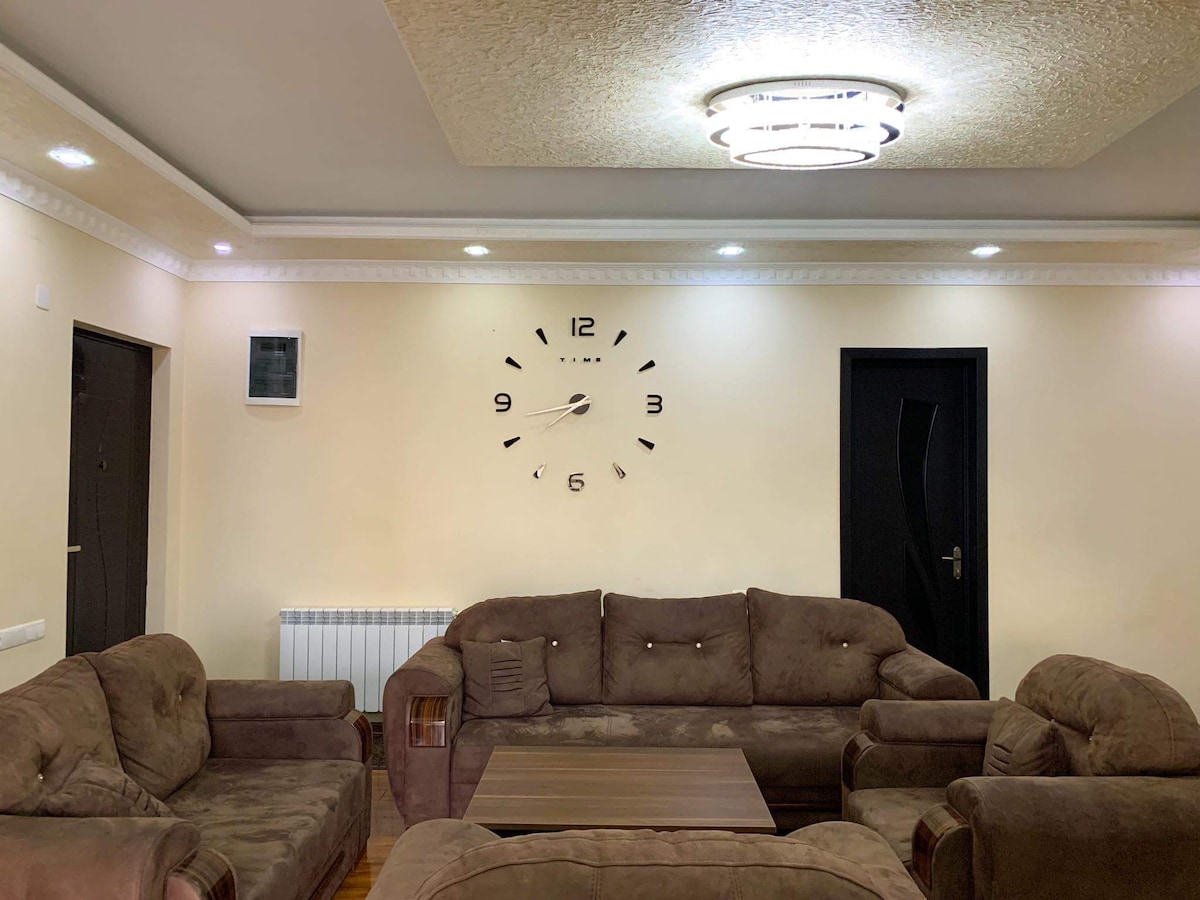 A spacious living area features a soft brown couch arrangement surrounding a low coffee table. A large wall clock is positioned above the sofa, while recessed lighting enhances the room’s warmth. A dark door is visible, leading to another area.