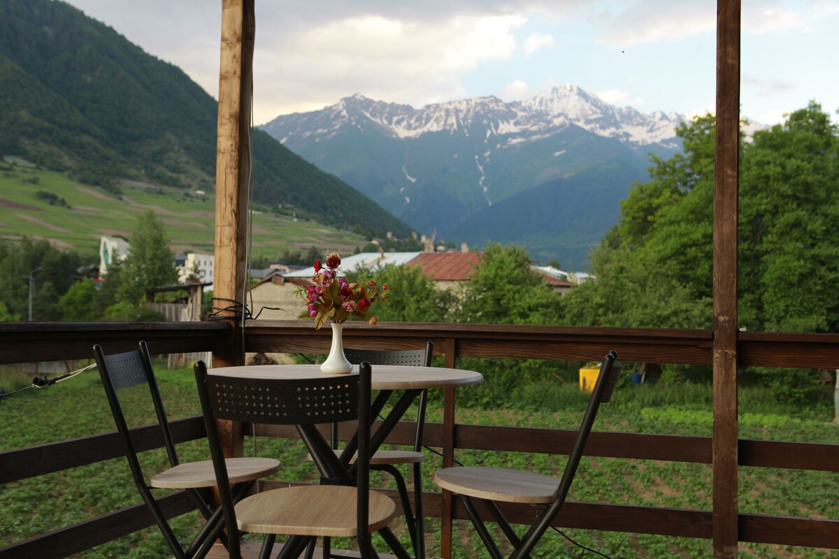 A wooden terrace features a round table surrounded by four chairs. A small vase of flowers is placed on the table. In the background, snow-capped mountains rise majestically, with greenery filling the foreground, creating a serene outdoor setting.