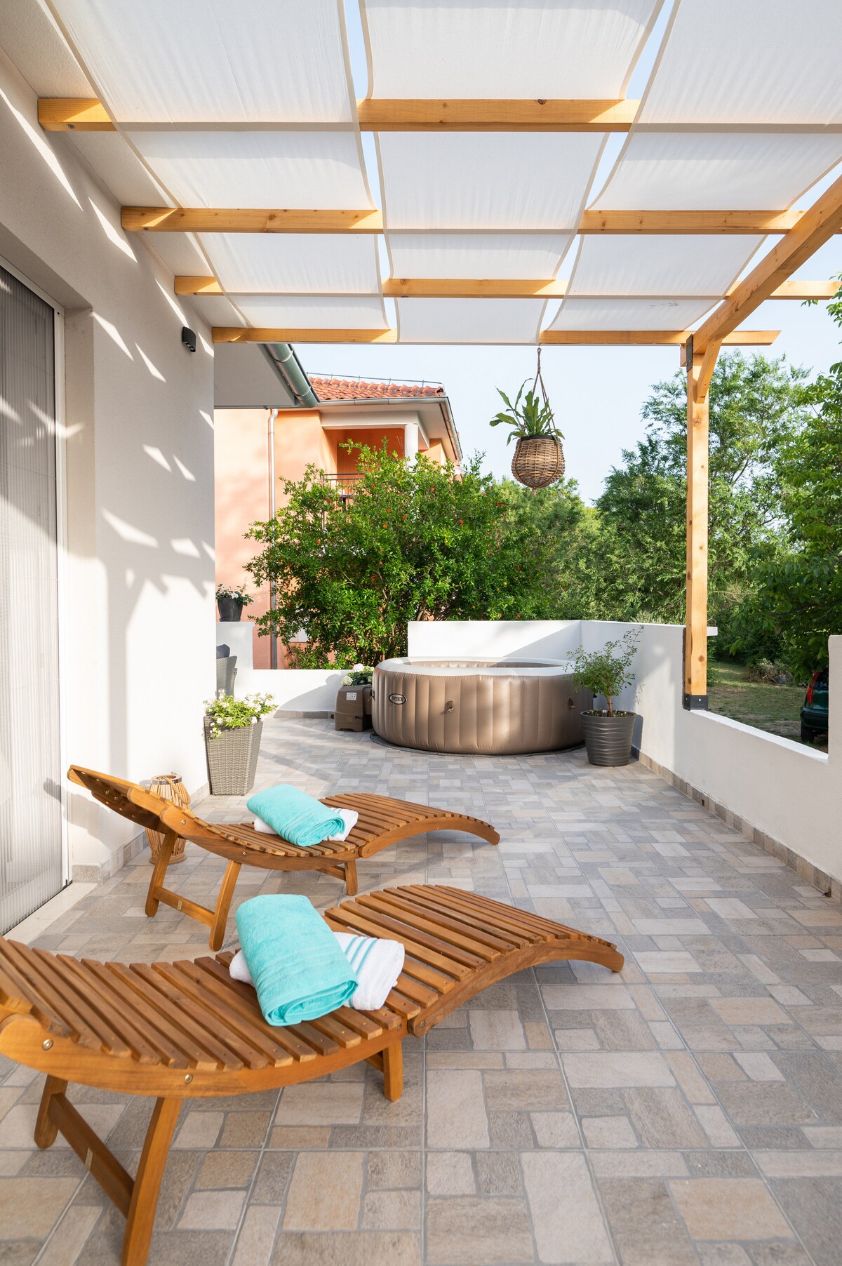 A spacious terrace is shown with wooden lounge chairs, accented by rolled towels. A jacuzzi is located in the background, surrounded by greenery. Natural light filters through a canopy overhead, creating a serene atmosphere, perfect for relaxation.