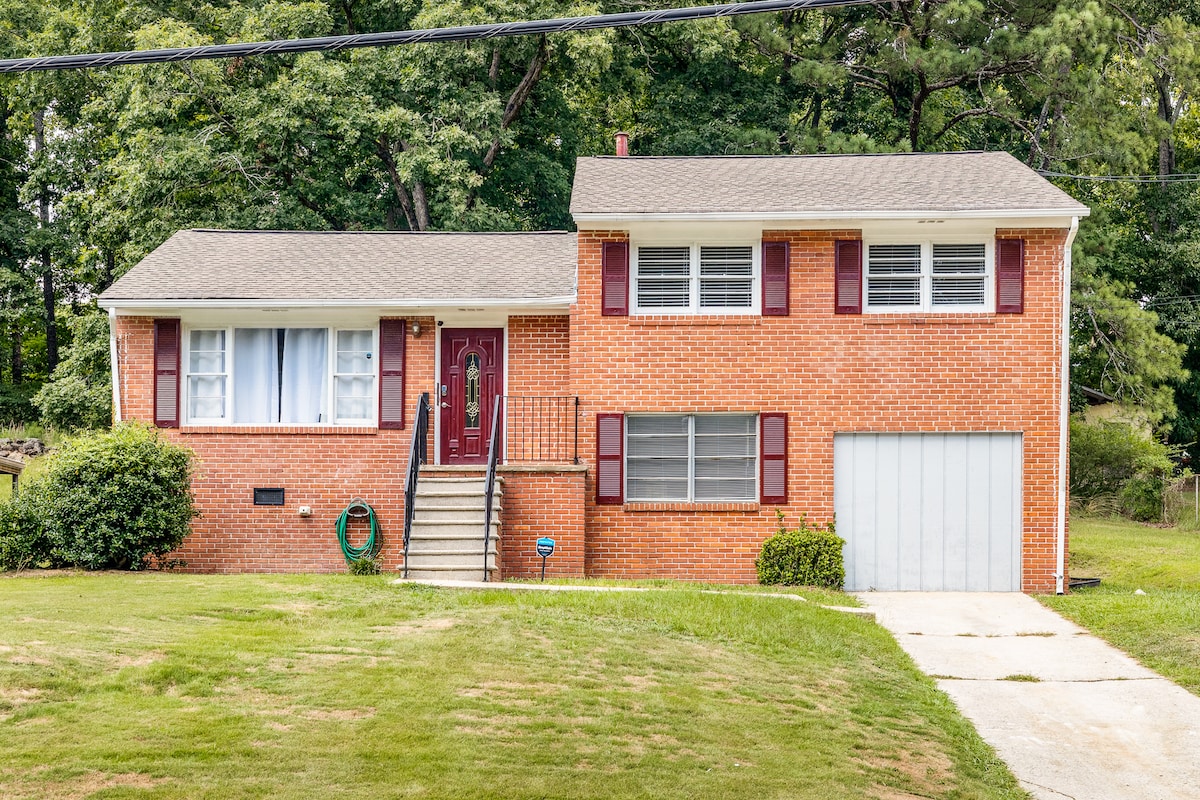 A two-story brick home is depicted, featuring a welcoming front entrance with a red door. Large windows are framed with maroon shutters. A well-manicured lawn surrounds the property, and a driveway leads to a garage on the right side.