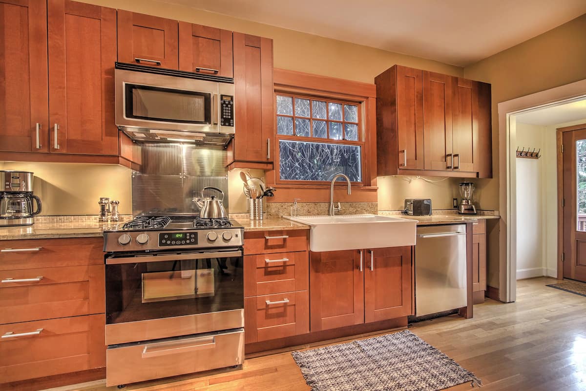 The kitchen features warm wooden cabinets and granite countertops. A stainless-steel stove and microwave are situated against the wall, complemented by a farmhouse sink. Modern appliances, including a dishwasher and coffee maker, are visible. Natural light filters through the window, enhancing the inviting atmosphere.