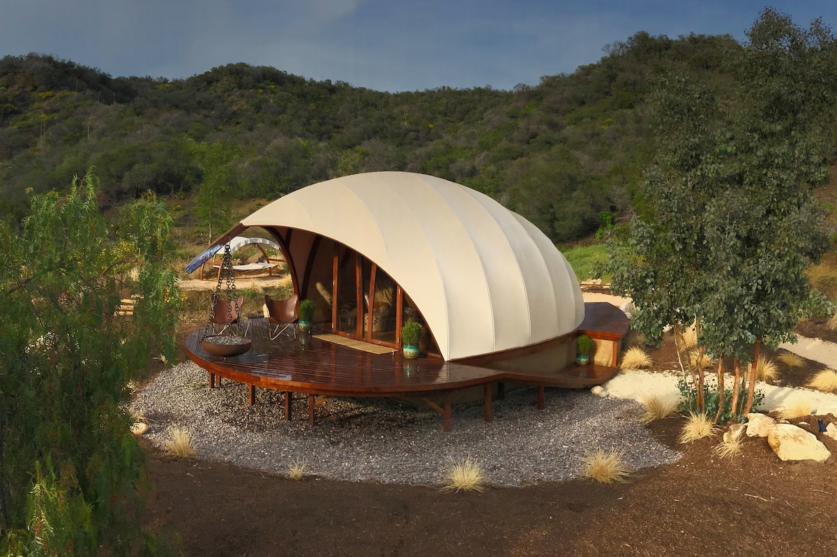The Gesner Cocoon is showcased with its distinctive architectural design featuring a curved, fabric roof. Wood decking surrounds the structure, complemented by natural landscaping and gravel. The backdrop includes rolling green hills, enhancing the secluded feel of the glamping site.