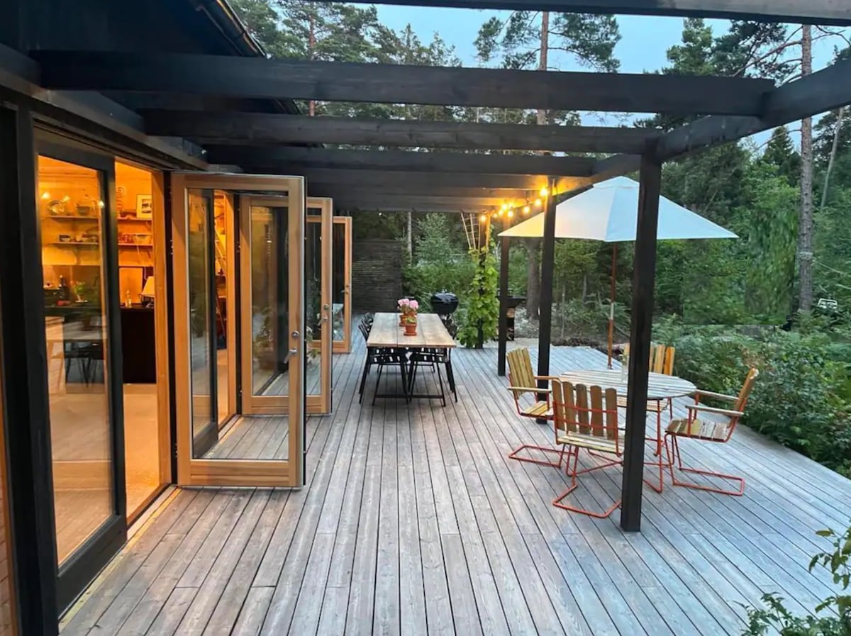 A spacious wooden deck is shown, featuring a long dining table and several chairs. Glass doors open to reveal the interior, enhancing the connection with nature. Soft lighting is provided by string lights overhead, and a large umbrella offers shade for outdoor seating.