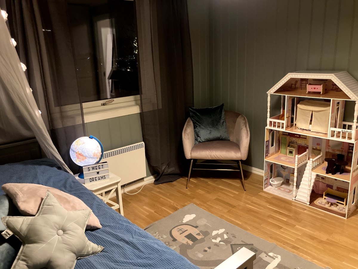A cozy bedroom features a spacious bed dressed in soft blue linens. A plush armchair sits beside a stylish dollhouse. A decorative globe and a rug with a whimsical design complement the ambiance, while dark curtains frame a window that offers nighttime views.