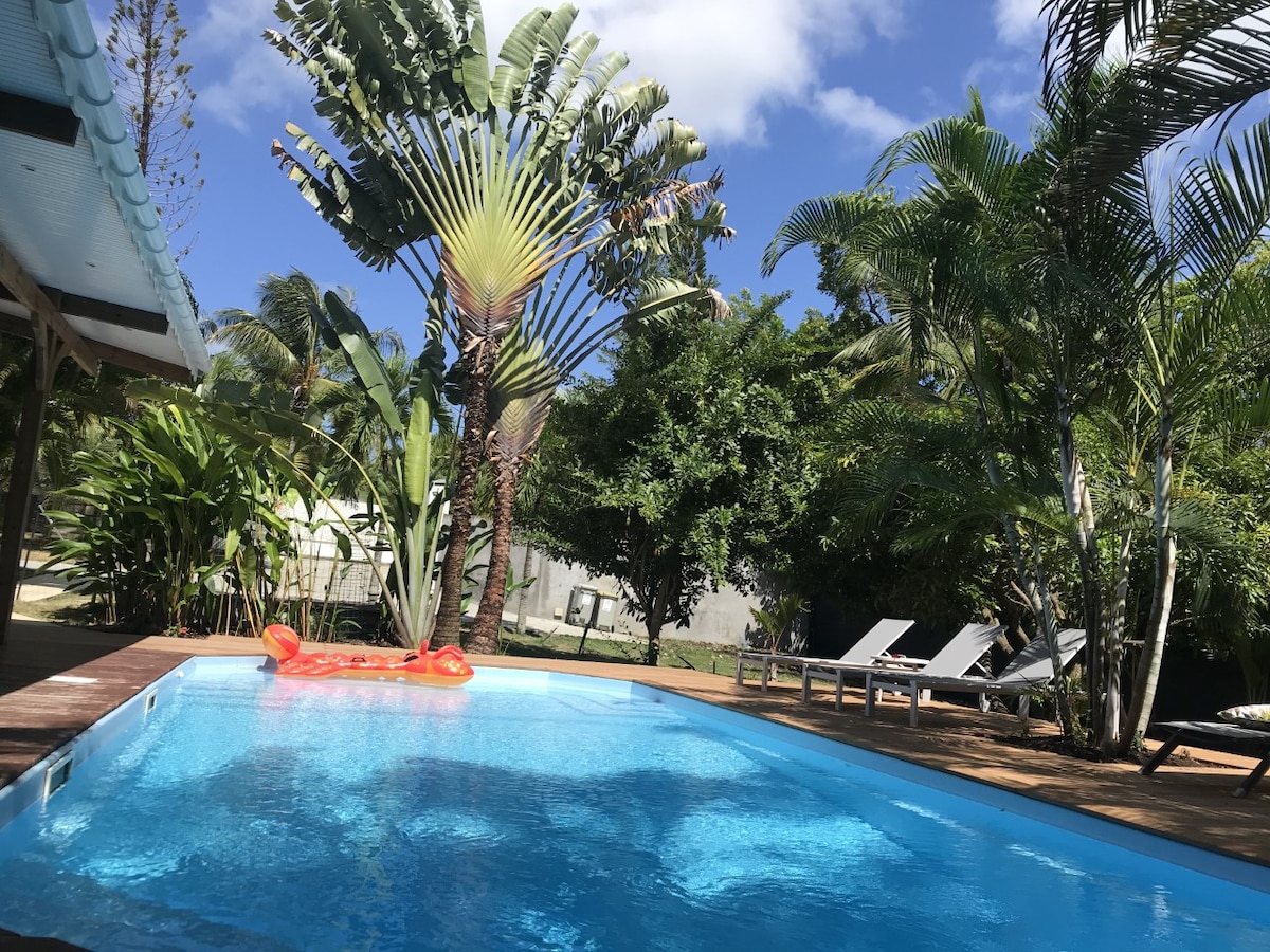 An inviting pool area is surrounded by lush greenery and palm trees. Lounge chairs are positioned on a wooden deck, and a bright orange float is seen resting on the clear blue water. The sky is clear, contributing to a serene outdoor setting.