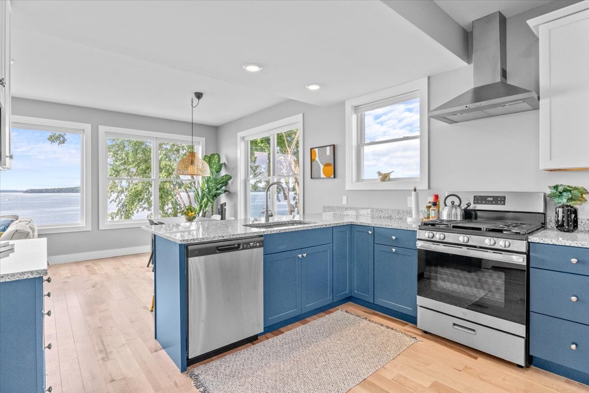 The open-concept kitchen is designed with blue cabinetry and granite countertops. Stainless steel appliances, including a dishwasher and oven, are visible. Large windows allow natural light to fill the space, with views of the lake visible in the background.