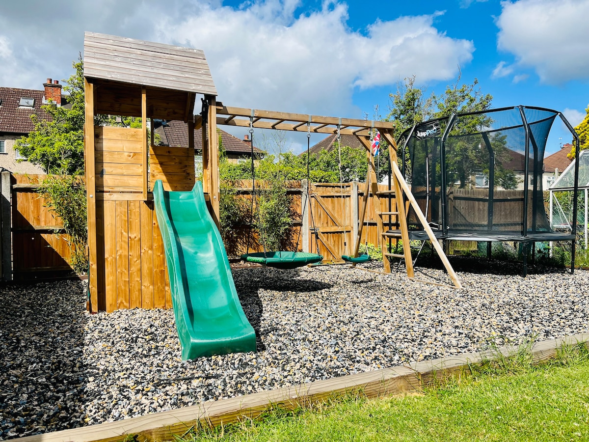 A children's play area features a wooden climbing fort with a green slide and swings. A trampoline with a safety net is positioned nearby, surrounded by a pebble surface and lush grass. The area is enclosed by wooden fencing and framed by greenery.