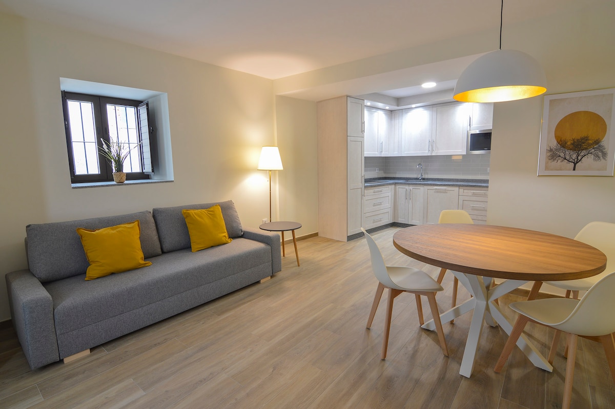 A light-filled living area is presented, featuring a gray sofa accented with yellow pillows, a wooden dining table surrounded by white chairs, and a floor lamp. A kitchenette with modern cabinetry is visible in the background, complemented by a window letting in natural light.