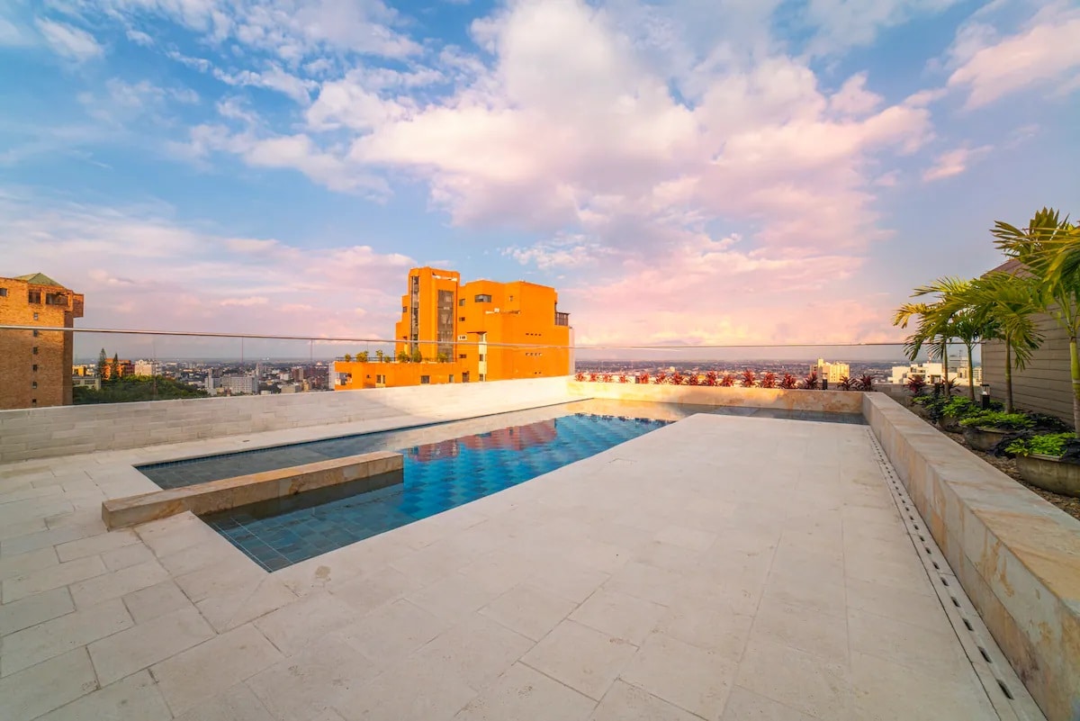 An outdoor pool area features a rectangular swimming pool set against a backdrop of a vibrant orange building. Surrounding the pool, a sun terrace is adorned with lounge chairs, offering a scenic view under a colorful sky with scattered clouds.