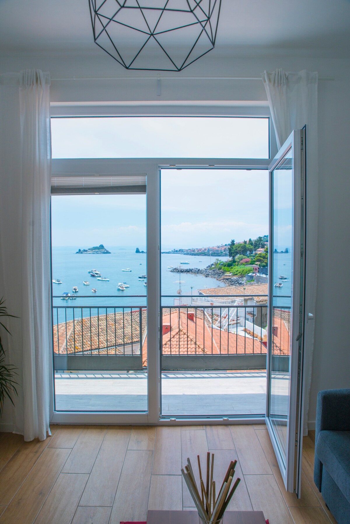 A large glass door opens to a balcony with a view of the sea, framed by light curtains. The landscape includes boats gently bobbing in the water and the hilly coastline in the background, providing a serene maritime atmosphere.