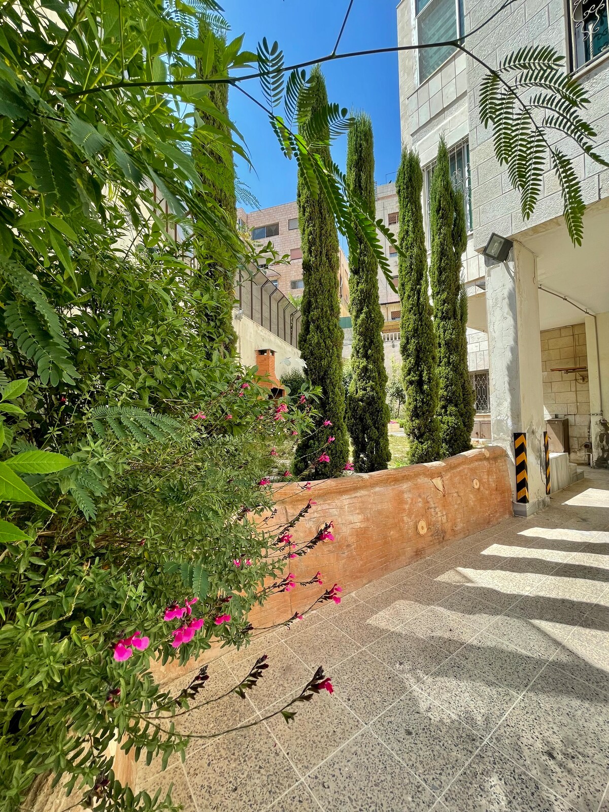 A serene outdoor area features tall cypress trees and blooming flowers, creating a peaceful atmosphere. Sunlight filters through the leaves, casting soft shadows on the stone pathway. The surrounding architecture is modern, adding a touch of elegance to the inviting space.