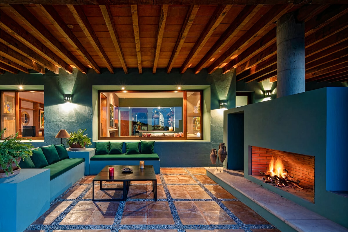 An inviting outdoor living area features a built-in fireplace with a cozy fire, surrounded by a stone-paved floor. Comfortable seating arranged along the walls provides a relaxed atmosphere, while large windows overlook a beautifully designed interior space.