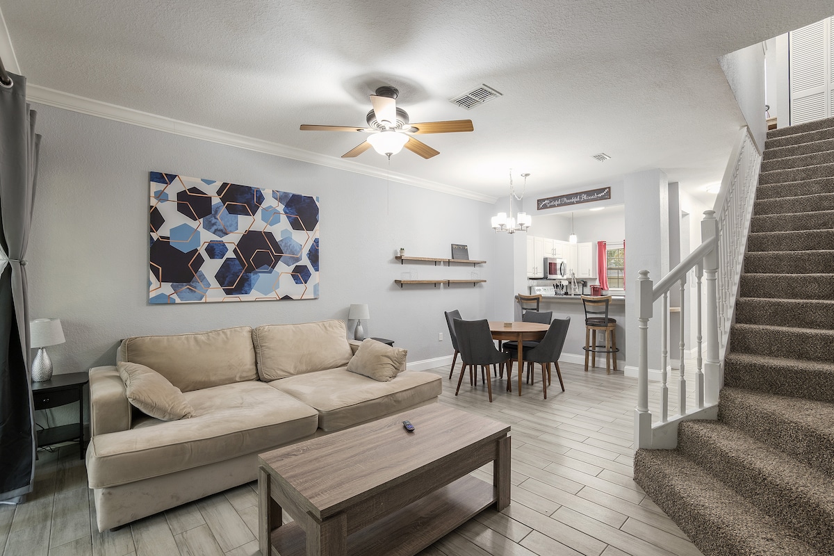 A spacious living and dining area features a comfortable sectional sofa, a coffee table, and a round dining table. Light wood flooring enhances the open space. A modern abstract art piece adorns the wall, with a ceiling fan providing additional comfort.