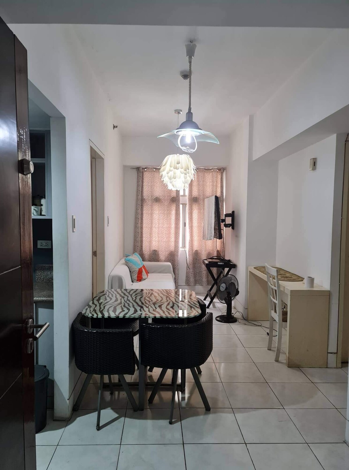 An inviting dining area features a glass-top table surrounded by dark wicker chairs. A cozy white sofa is positioned nearby, complemented by a colorful cushion. Light filters through sheer curtains, softly illuminating the space. A stylish pendant light hangs overhead.
