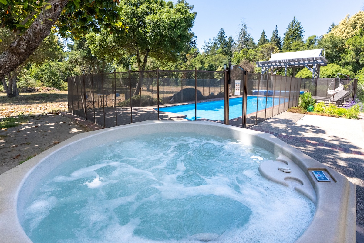 A hot tub is positioned beside a fenced pool area, with water bubbling gently. The pool can be seen in the background, surrounded by trees and greenery. A clear blue sky complements the outdoor setting.