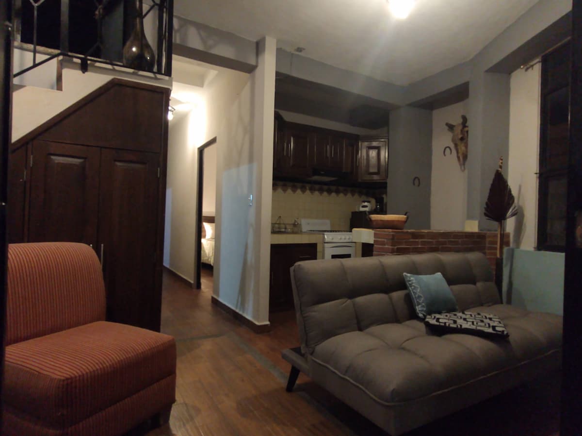 An inviting living area features a gray sofa bed facing a small balcony. A bright armchair adds a pop of color, while the kitchen area is partially visible, showcasing wooden cabinetry. Stairs lead to a second level, and soft overhead lighting creates a warm ambiance.