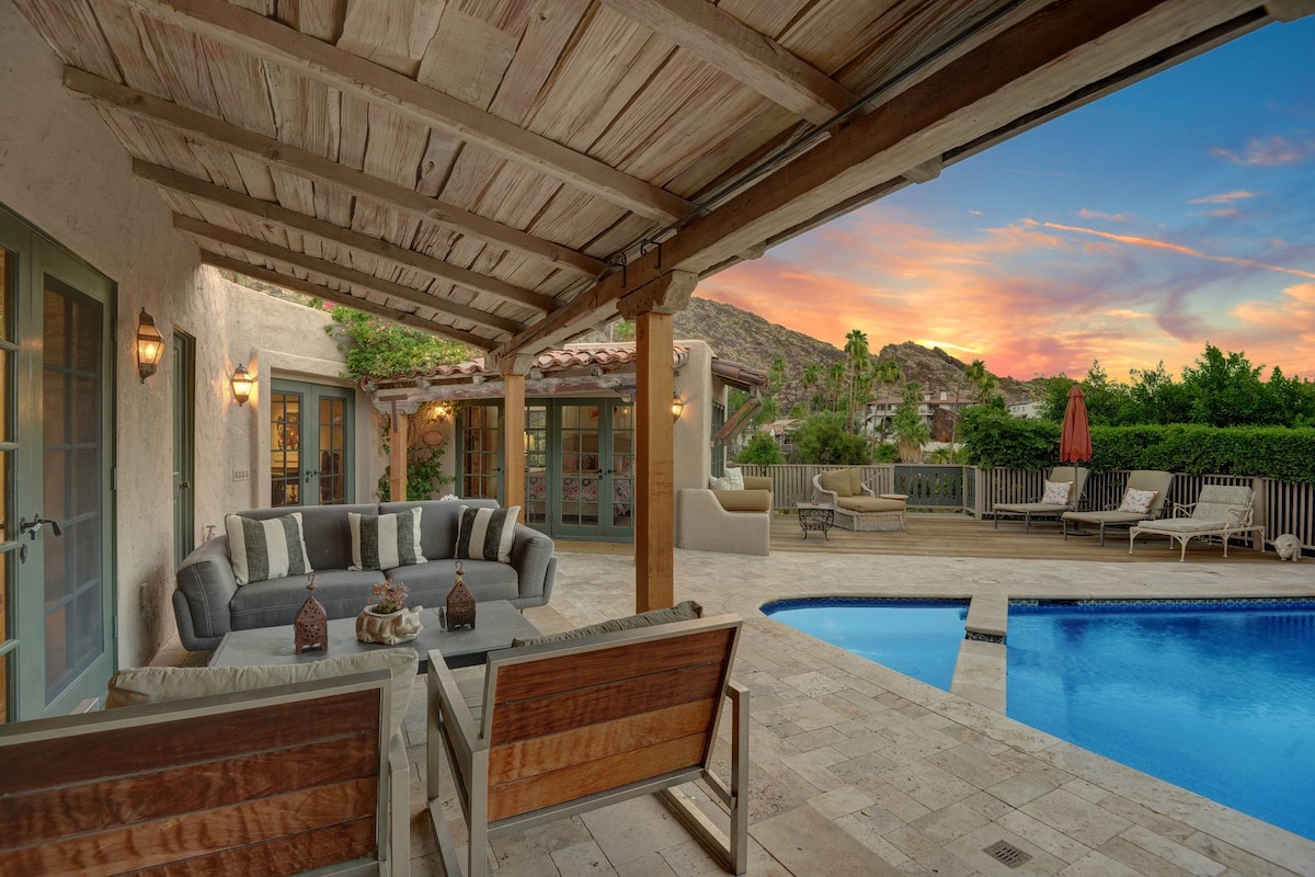 An outdoor living area is showcased with comfortable seating arranged around a stone table. A pool glimmers in the foreground, while a spacious sundeck is seen in the background with lounge seating and umbrellas. Lush greenery frames the area, complementing the serene desert landscape.