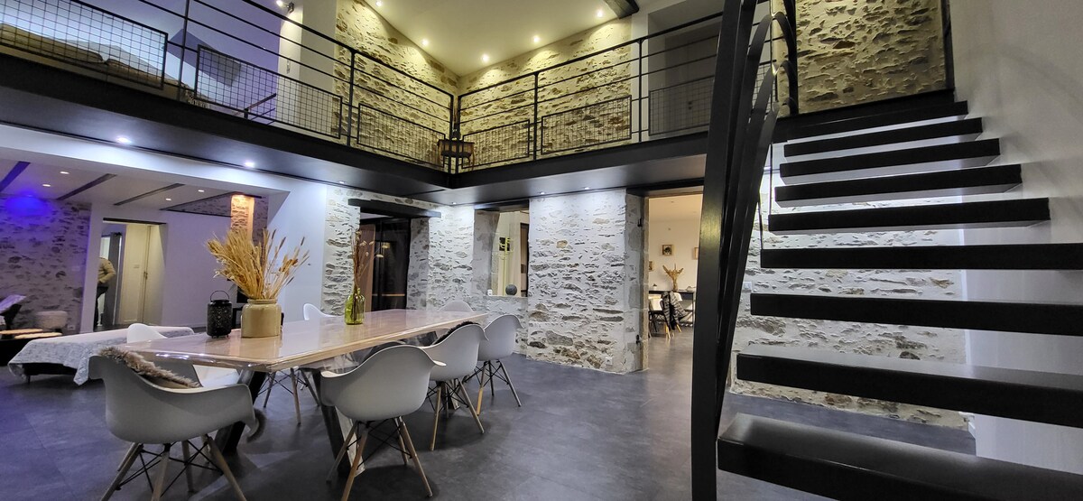 The spacious dining area features a long wooden table surrounded by modern chairs. Warm lighting highlights the stone walls, while a spiral staircase leads to an upper level. The open concept layout creates a seamless integration of the living space and dining area.