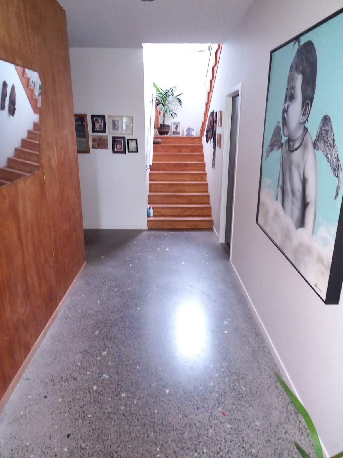 A corridor is seen with polished concrete flooring, accented by a large art piece depicting a child with wings on the right wall. Photographs are displayed on the left wall, and a staircase with wooden steps leads to the upper level, illuminated by natural light.