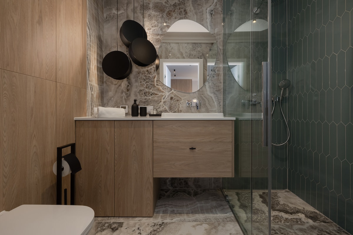 A modern bathroom features a combination of textured wood cabinetry and striking tiled walls. Sleek mirrors and circular accents enhance the design, while a walk-in shower is visible behind clear glass. A neutral color palette contributes to a clean and inviting environment.