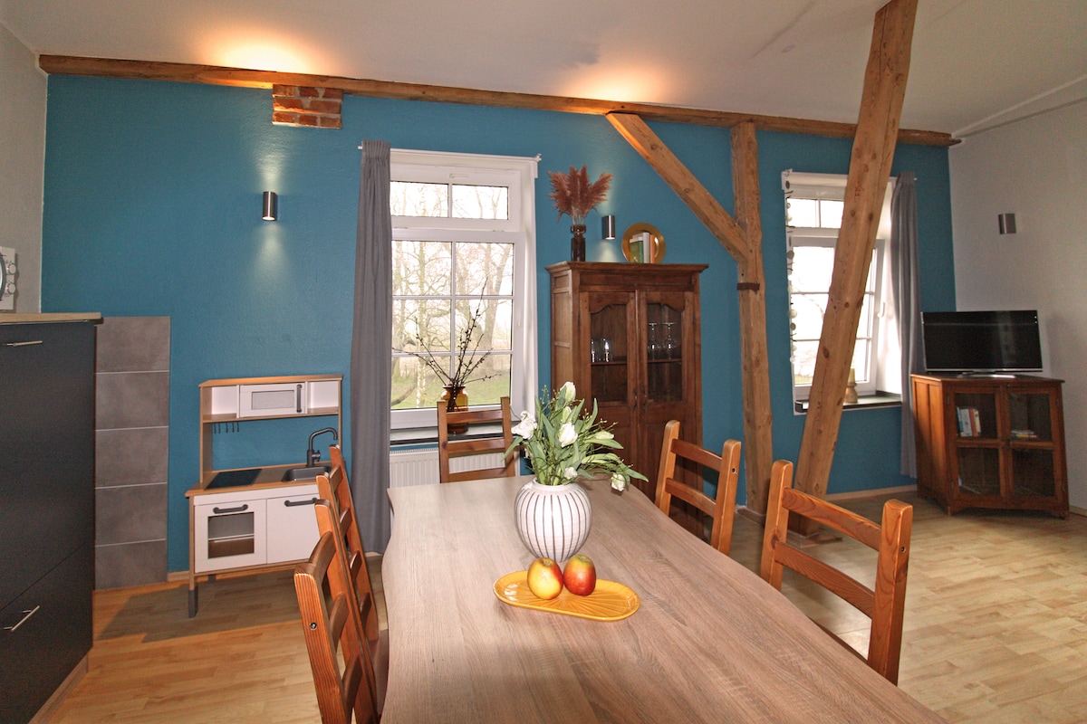 A welcoming dining area features a wooden table surrounded by chairs, illuminated by natural light from the large windows. A decorative wooden cabinet and a small kitchenette are present, with the walls painted in a calming blue hue and exposed wooden beams adding character.