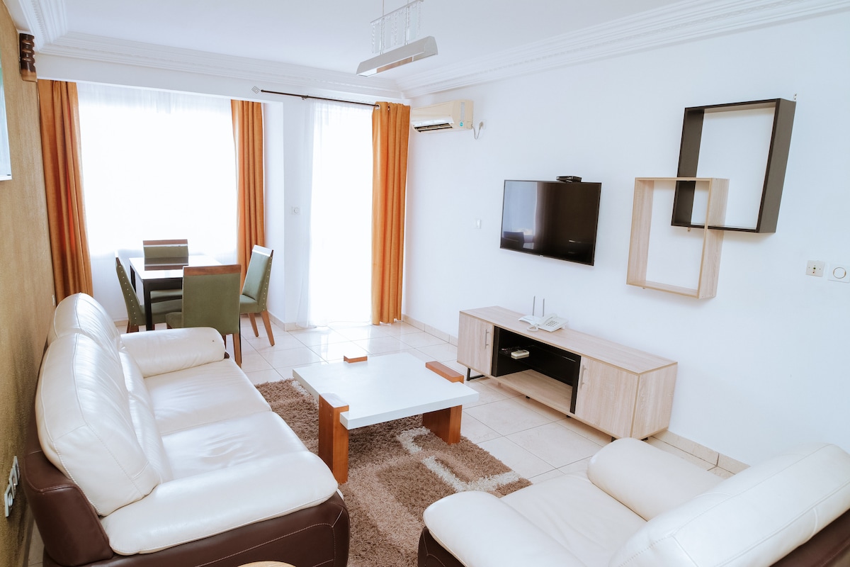 A bright living area features a comfortable seating arrangement with white leather sofas positioned around a central coffee table. A flat-screen television is mounted on the wall, while a dining table is visible in the background, complemented by green chairs and natural light streaming through large windows.
