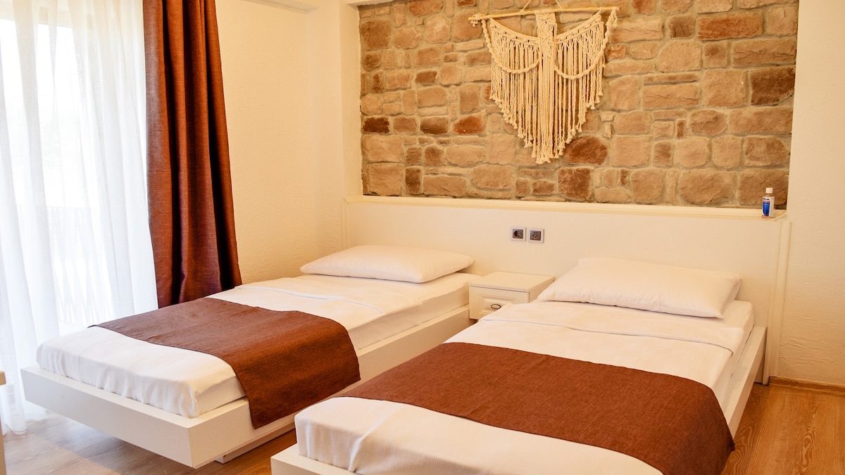 The bedroom features two spacious beds with crisp white linens and brown throws. A natural stone wall adds texture, while a decorative wall hanging complements the design. Soft, sheer curtains allow light to filter through, creating a calm ambiance.