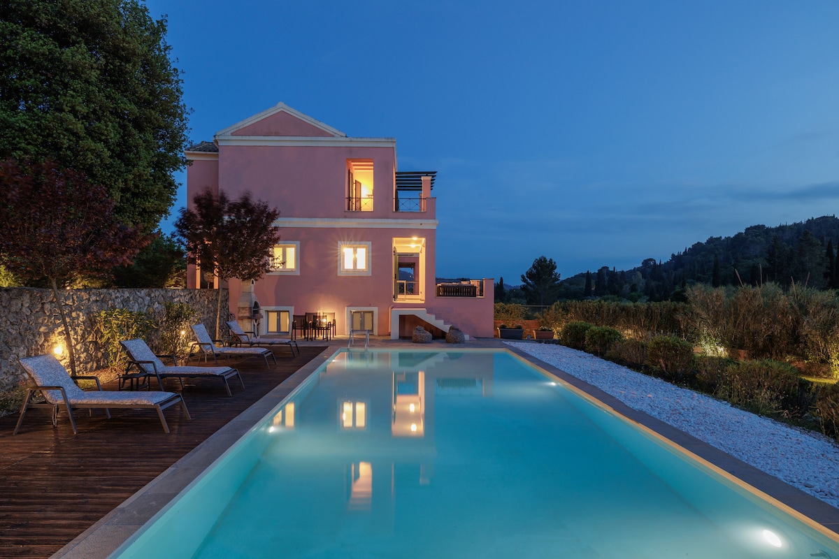 The villa, featuring a charming pink exterior, is illuminated softly at dusk. A private swimming pool reflects the evening sky, surrounded by lounge chairs on a wooden deck and a landscaped garden that adds to the serene setting.
