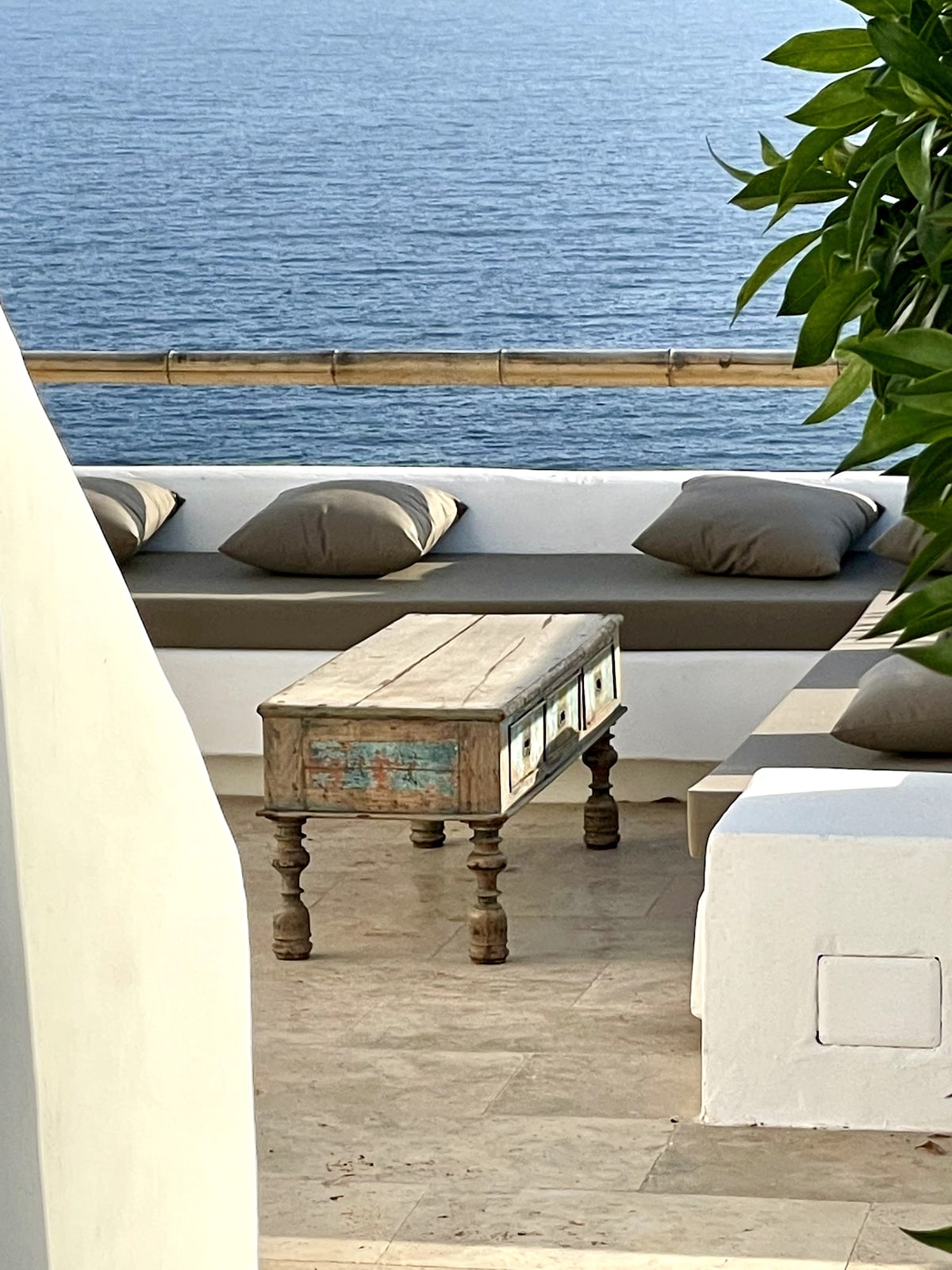 A cozy outdoor seating area overlooks the sea, featuring a low table with an antique finish and several plush cushions arranged on the seating. The space is surrounded by greenery, providing a serene environment for relaxation.