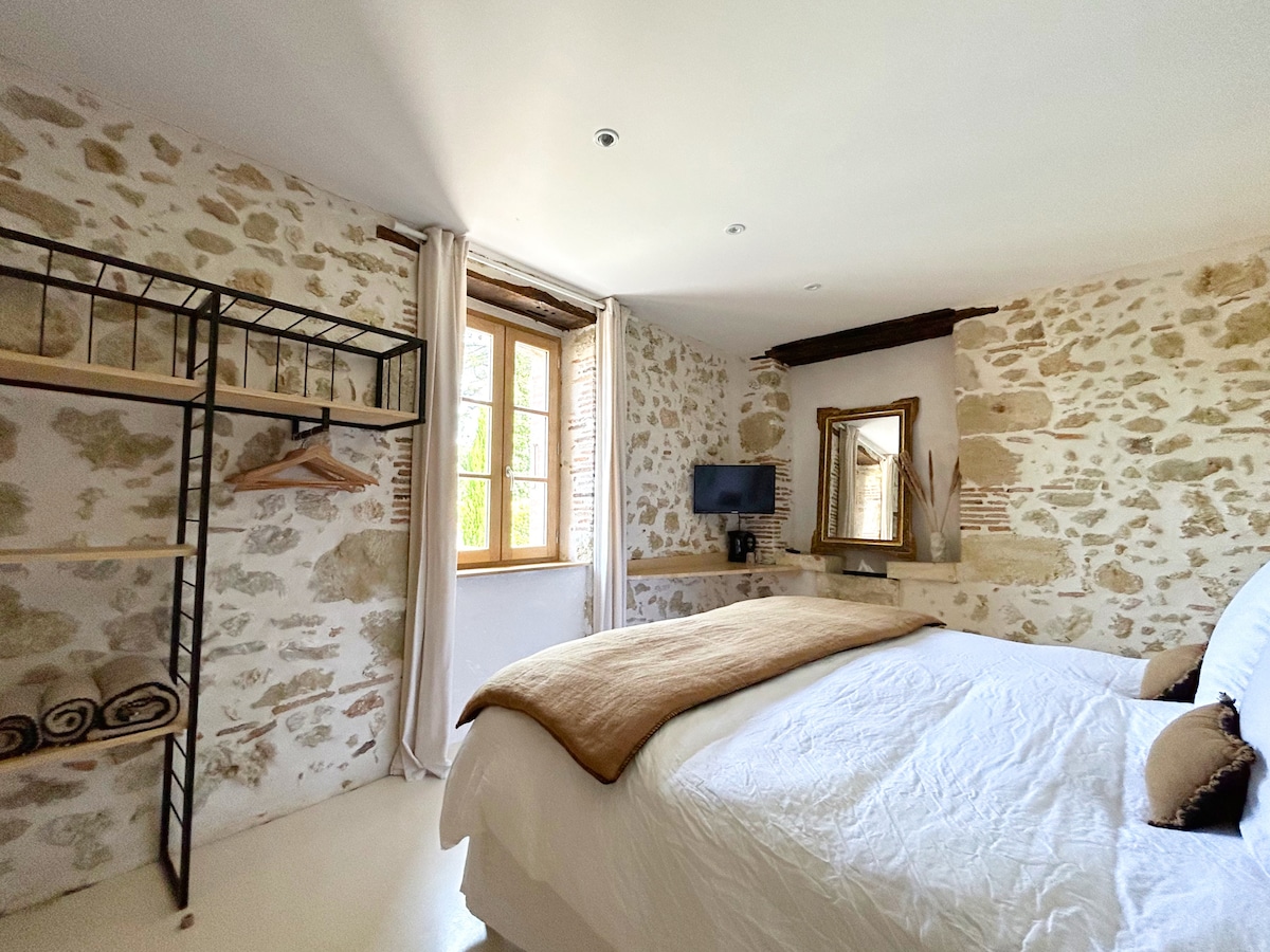 A serene bedroom features exposed stone walls and softly lit windows. The spacious bed is adorned with neutral linens and accompanied by a wooden frame mirror. A small television is mounted on the wall, and open shelving showcases neatly arranged towels.