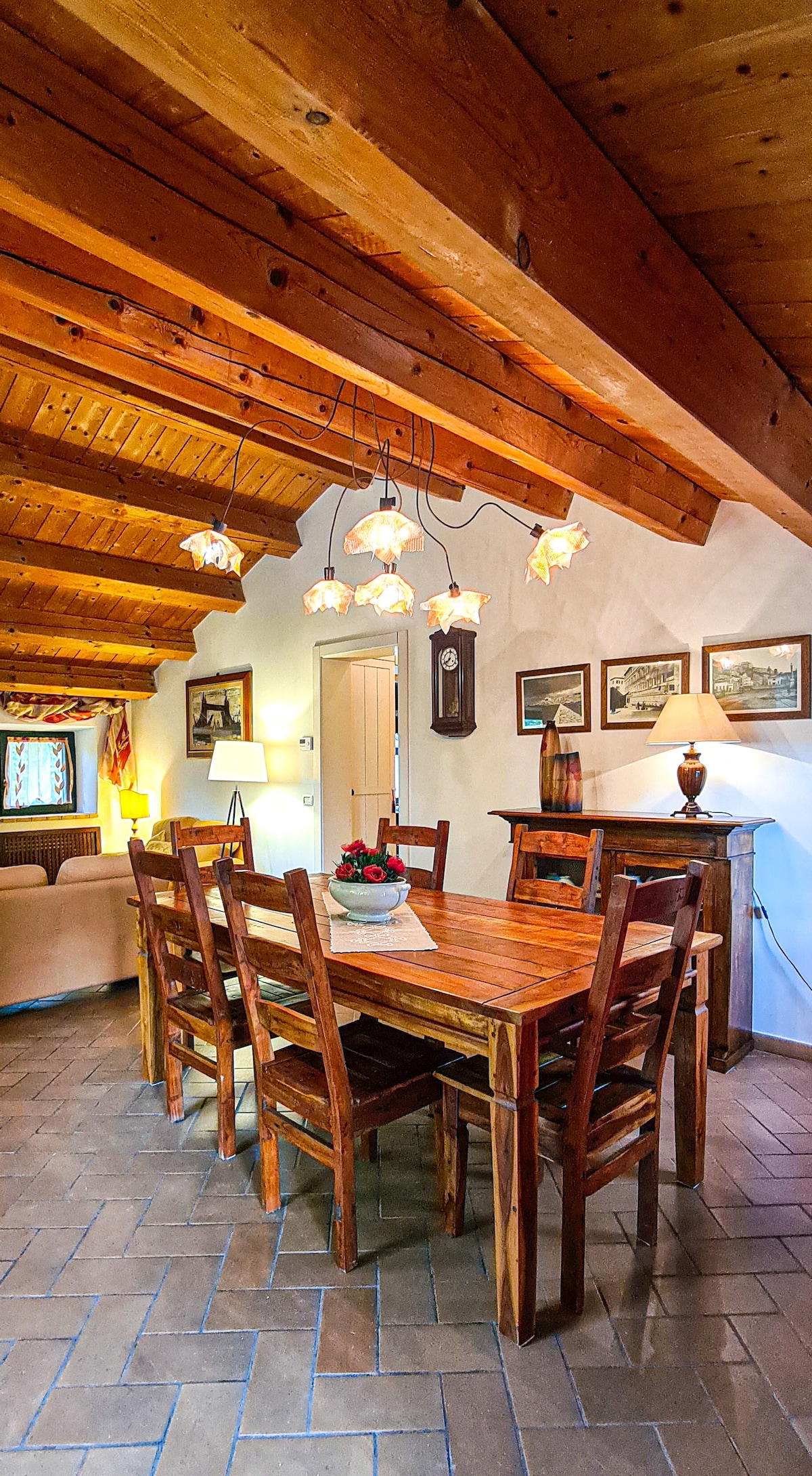 A dining area is illuminated by a unique light fixture, featuring floral designs. A wooden table is surrounded by sturdy chairs, with framed artwork adorning the walls. The ceiling showcases exposed wooden beams, contributing to the rustic charm of the space.