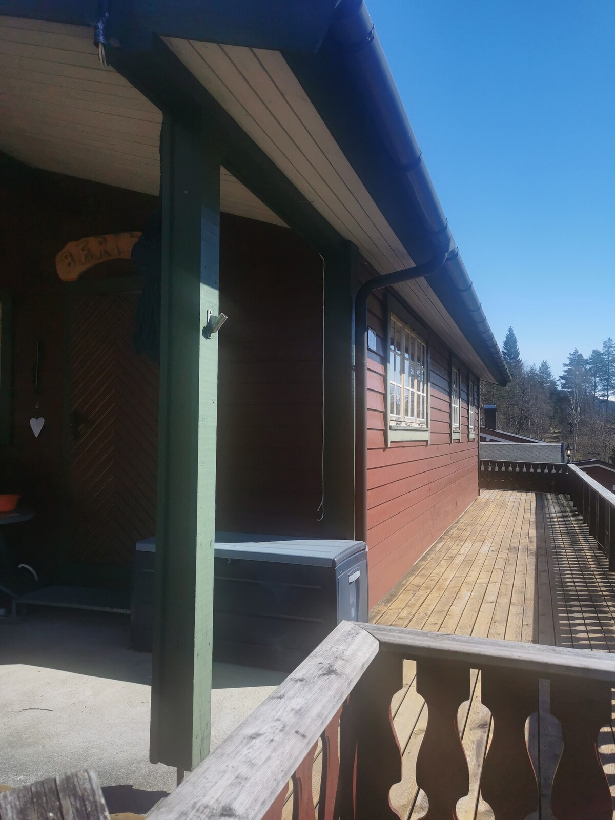 A wooden deck extends alongside the home, showcasing a warm red exterior. Large windows allow natural light to illuminate the interior, while a sturdy railing lines the deck. A storage box is positioned near the entrance, contributing to the functional outdoor space.
