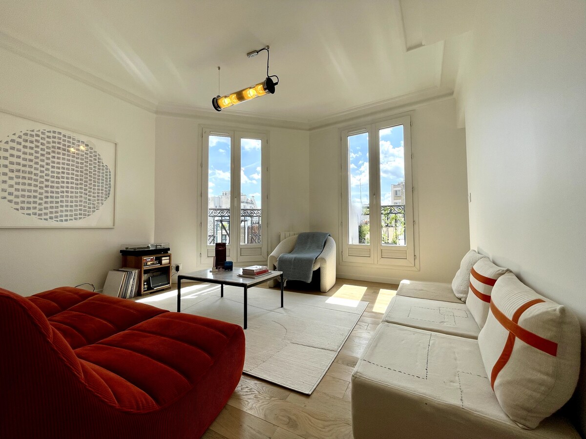 A bright living area features three distinct seating options, including a curved red sofa and a white upholstered sofa. A low black coffee table occupies the center space. The room is illuminated by natural light streaming through large windows, with a view of the skyline visible.