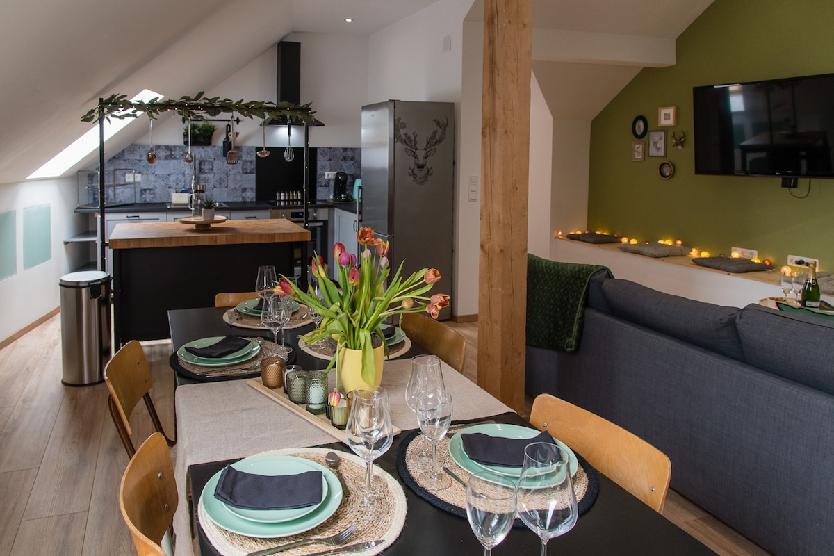 An open-plan space features a dining table set for six, adorned with vibrant flowers. A modern kitchen is highlighted by stainless steel appliances and an island with bar seating. Soft lighting adds warmth to the lounge area with a comfortable sofa and television.