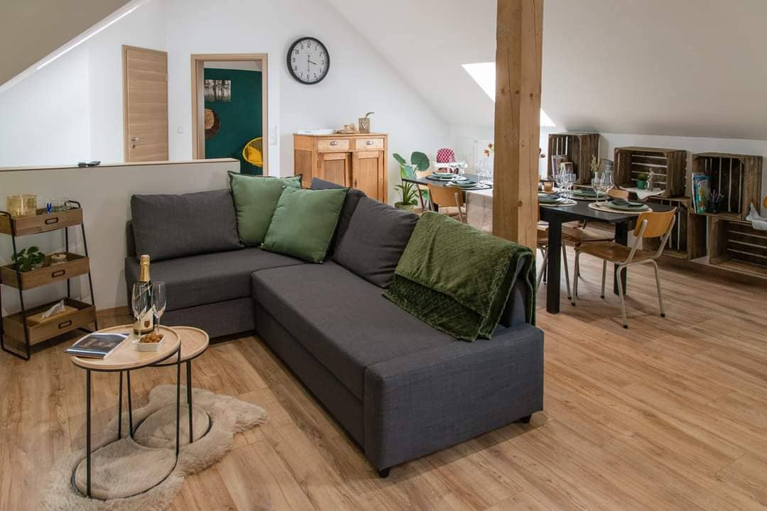 A spacious living area features a dark gray sectional sofa adorned with green throw pillows. A small round table holds a bottle of sparkling wine and glasses. In the background, a dining table is set with tableware, and natural light streams through angled roof windows.