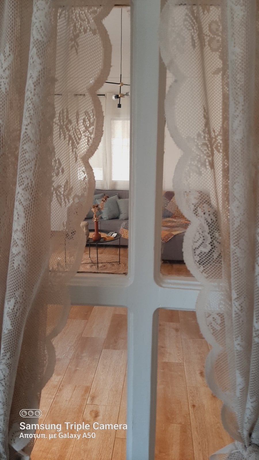 A delicate lace curtain frames a view through a window, leading to a tastefully furnished living area. Subtle lighting highlights a gray sofa, complemented by decorative cushions and a coffee table in the distance, creating an inviting and cozy ambiance.