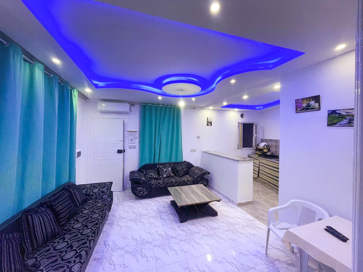 The living area features a modern design with black sofas arranged around a central coffee table. Soft blue lighting accents the ceiling, complemented by turquoise curtains that frame a window. A kitchen area is visible in the background, equipped with essential amenities.