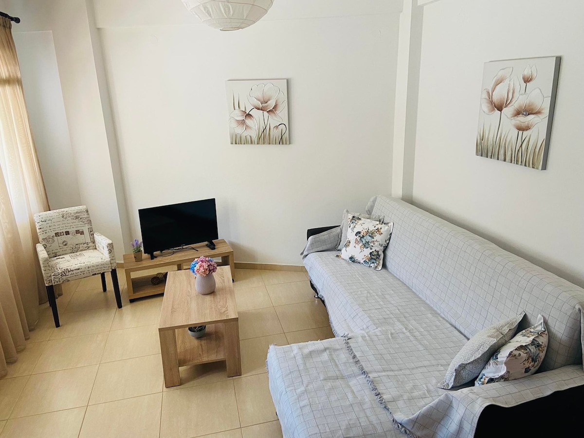 A comfortable living area features a gray couch and a wooden coffee table adorned with a floral arrangement. A wall-mounted TV is positioned opposite the seating. Two decorative wall art pieces enhance the space, accompanied by a chair with a patterned upholstery near a window with flowing curtains.