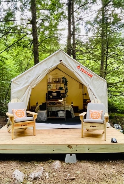 A spacious canvas tent sits on a wooden deck surrounded by lush greenery. Two Adirondack chairs, each with decorative cushions, are positioned in front of the tent entrance, inviting relaxation. Inside the tent, various camping essentials are neatly arranged for convenience.