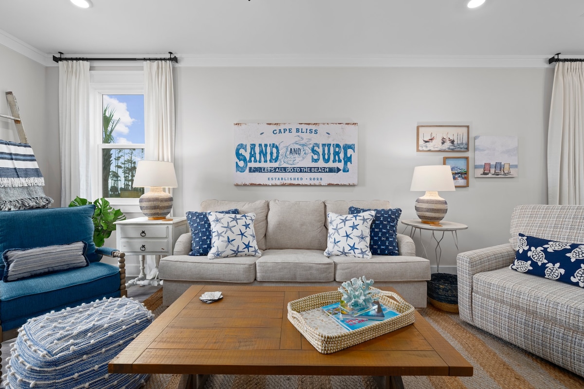 A welcoming living area features a light-colored sofa adorned with blue and white throw pillows. Two accent chairs in complementary shades provide additional seating. A wooden coffee table is positioned at the center, topped with decorative items. Artwork reflecting coastal themes is displayed on the walls.