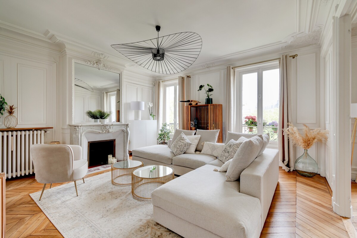 A spacious living area is present, featuring a light-colored sectional sofa and a round coffee table. Large windows allow natural light to fill the room, while elegant moldings adorn the walls. A decorative mirror and potted plants enhance the overall ambiance.