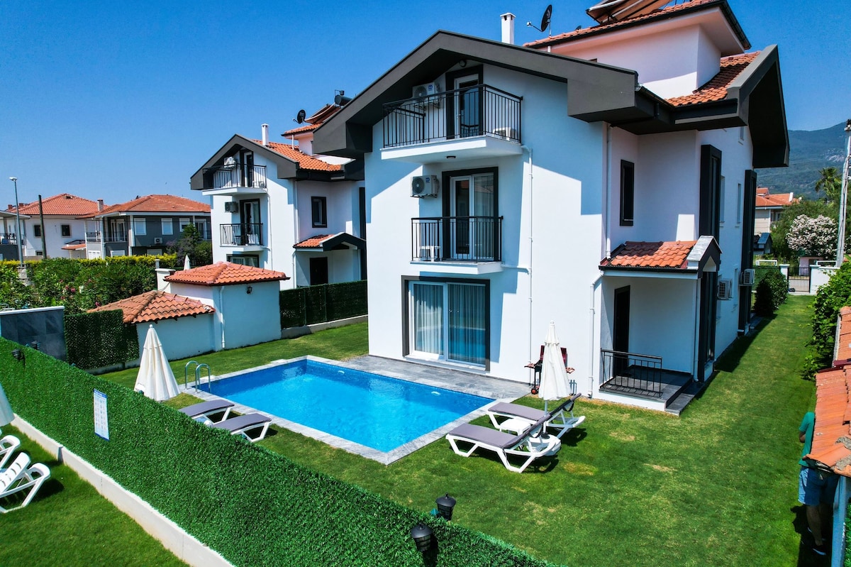 A modern two-story villa features a private pool surrounded by lush green grass. Lounge chairs are positioned beside the pool, and balconies are visible on the upper level. The exterior showcases a blend of white walls and red-tiled roofs, offering a spacious outdoor area.