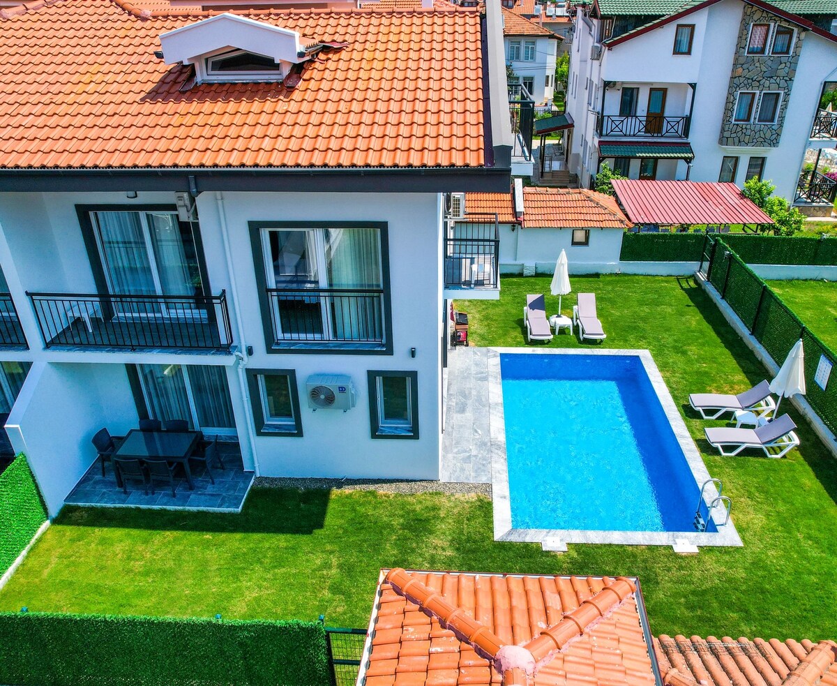 An aerial view captures a modern two-story villa surrounded by a well-maintained lawn. A sparkling blue pool is situated adjacent to the house, complemented by several lounge chairs and patio umbrellas. The roof features traditional tiles, adding a rustic charm to the property.